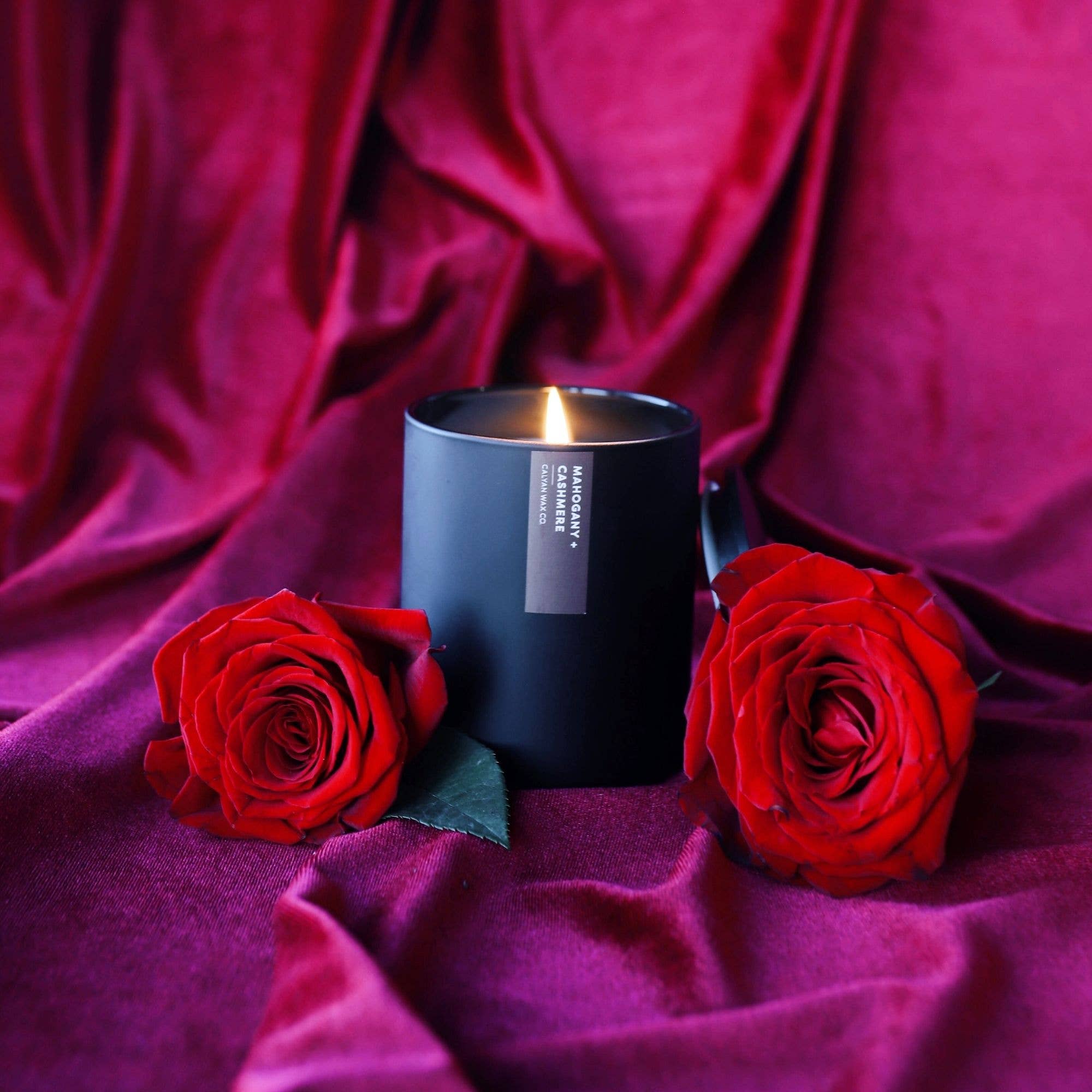Black candle with a label between two red roses on a purple fabric background