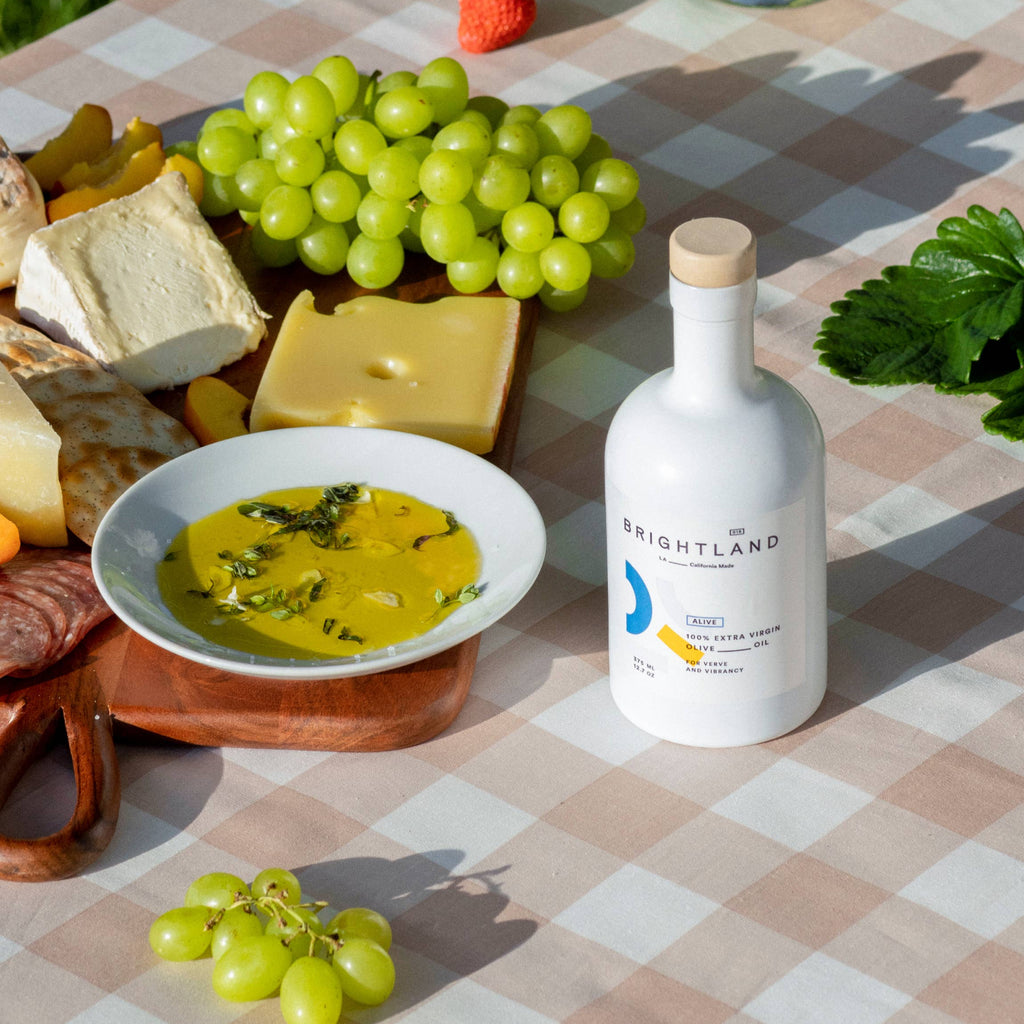 Bottle of Brightland olive oil on a picnic table with cheese, grapes, and other snacks.