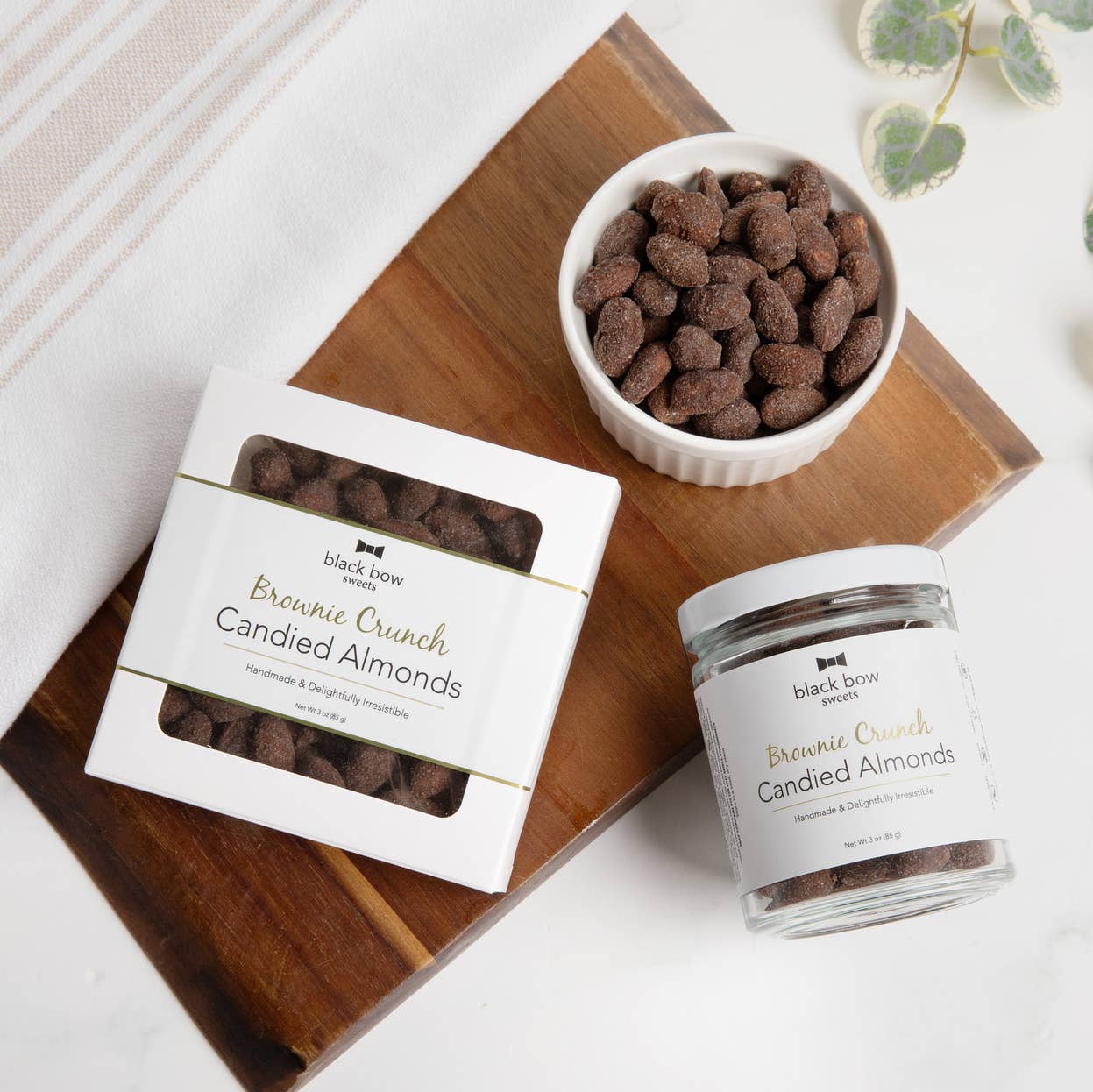 Bowl, box, and jar of 'Brownie Crunch Candied Almonds' on a wooden board.