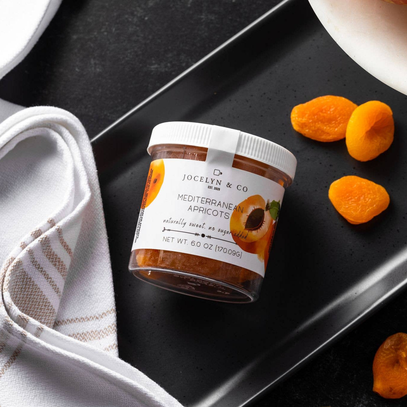 Jar of Jocelyn & Co. Mediterranean Apricots on a dark surface with apricots around