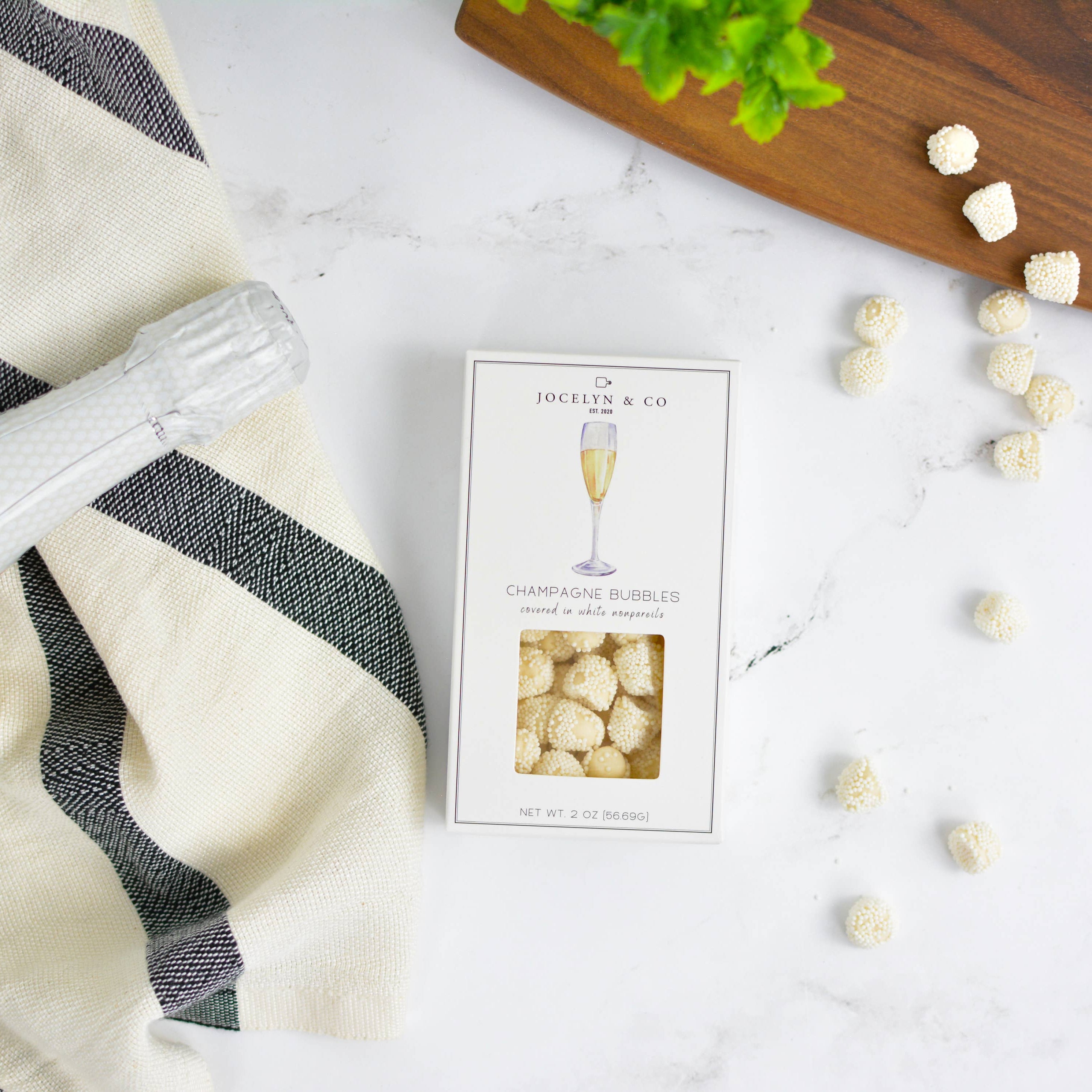 Bottle of champagne, a box of bubbles candy on a white surface with a black and white striped towel.