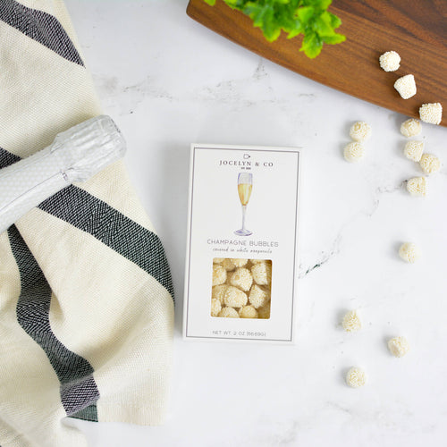 Bottle of champagne, a box of bubbles candy on a white surface with a black and white striped towel.