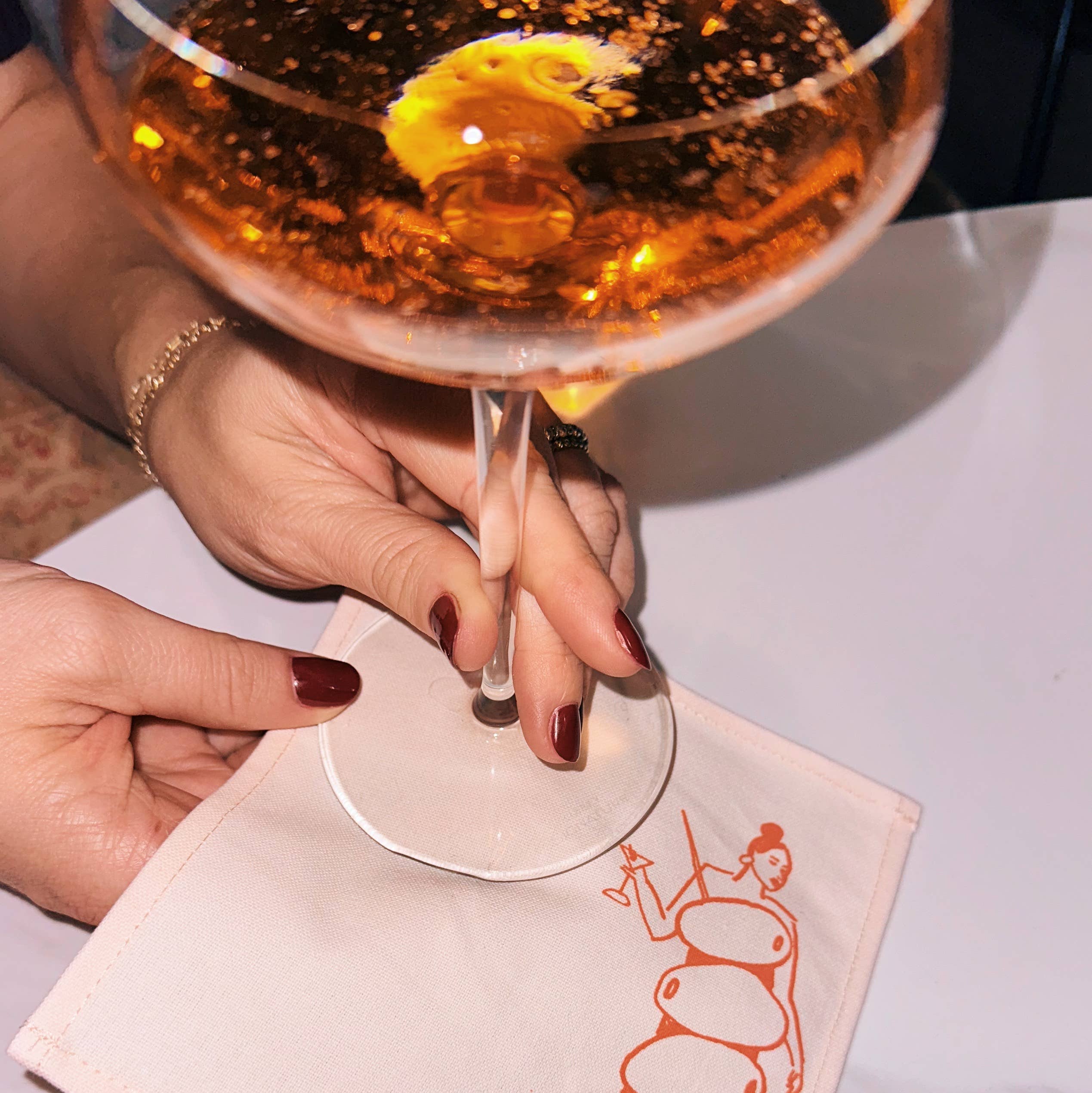 Hand holding a glass of amber liquid with a decorative napkin featuring a stylized figure on a white surface.