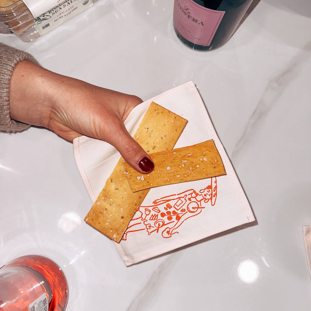 Hand holding a cracker with a decorative napkin on a white surface with bottles in the background