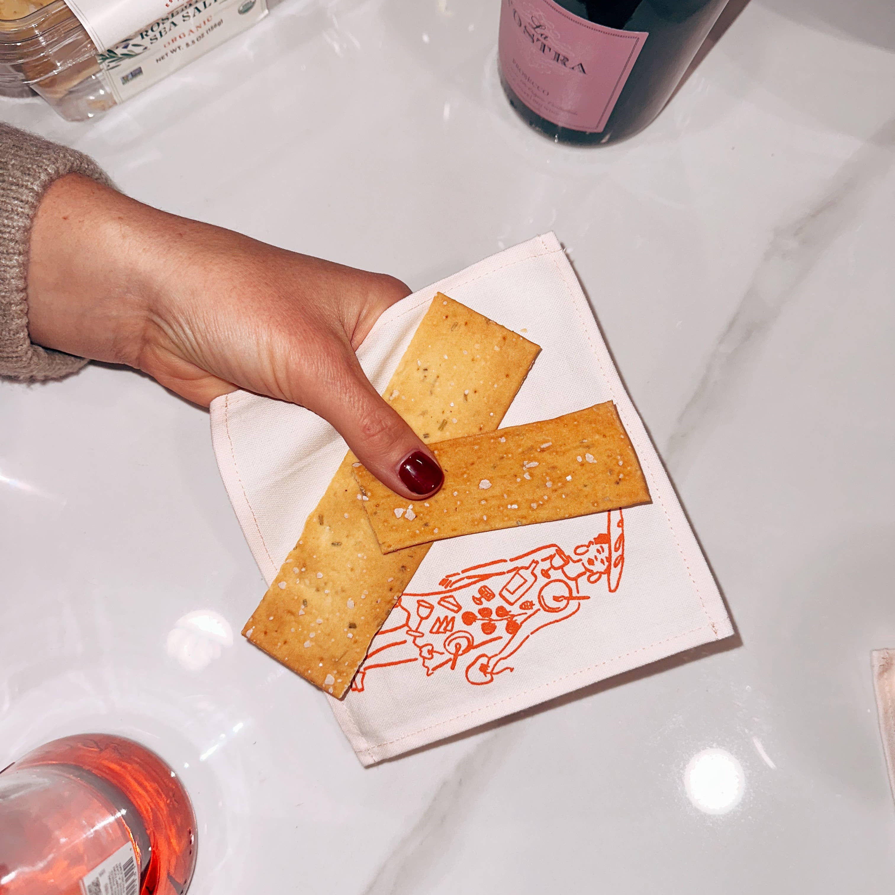 Hand holding a cracker with a decorative napkin on a white surface with bottles in the background