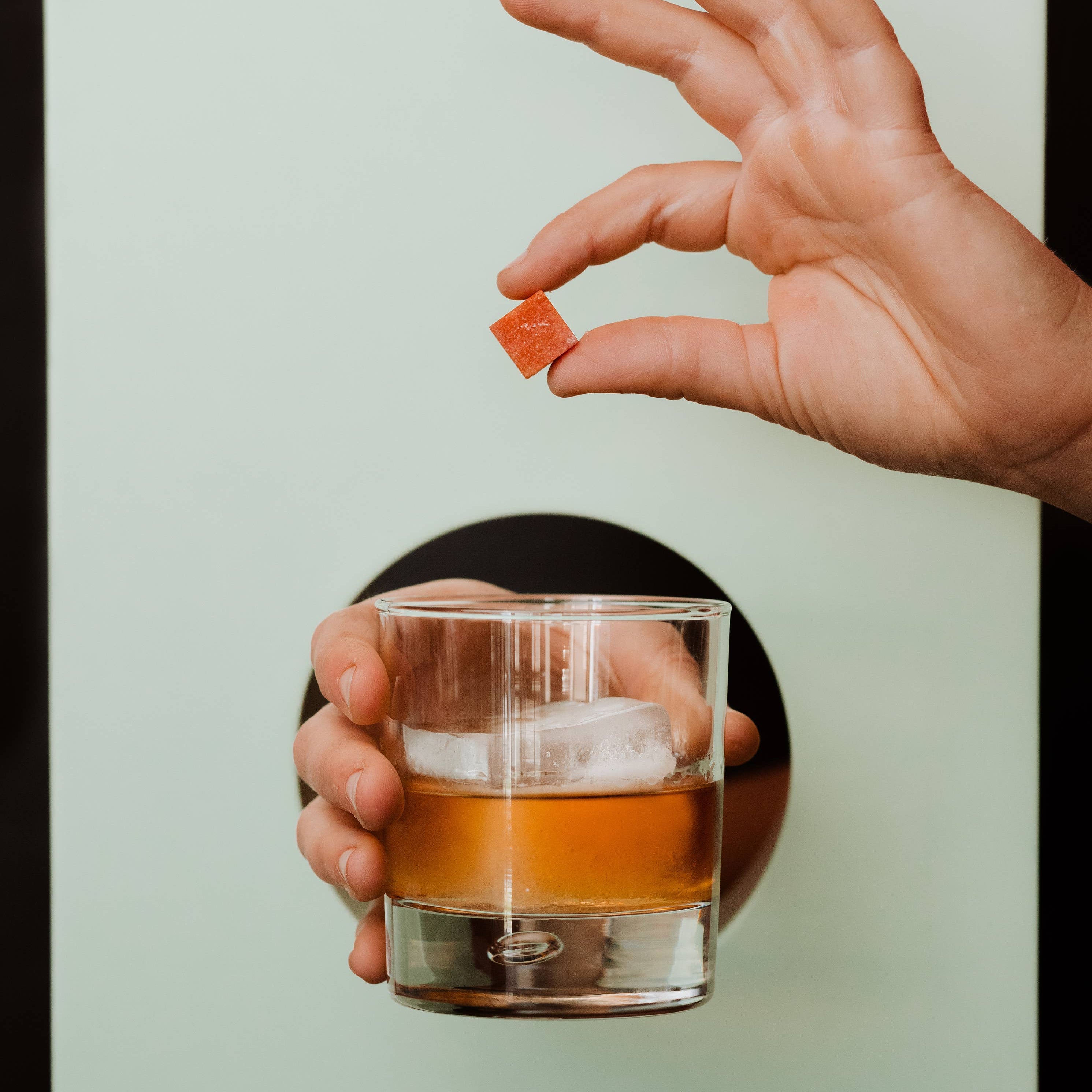 Hand holding a glass of amber liquid with a small orange candy, against a light green background