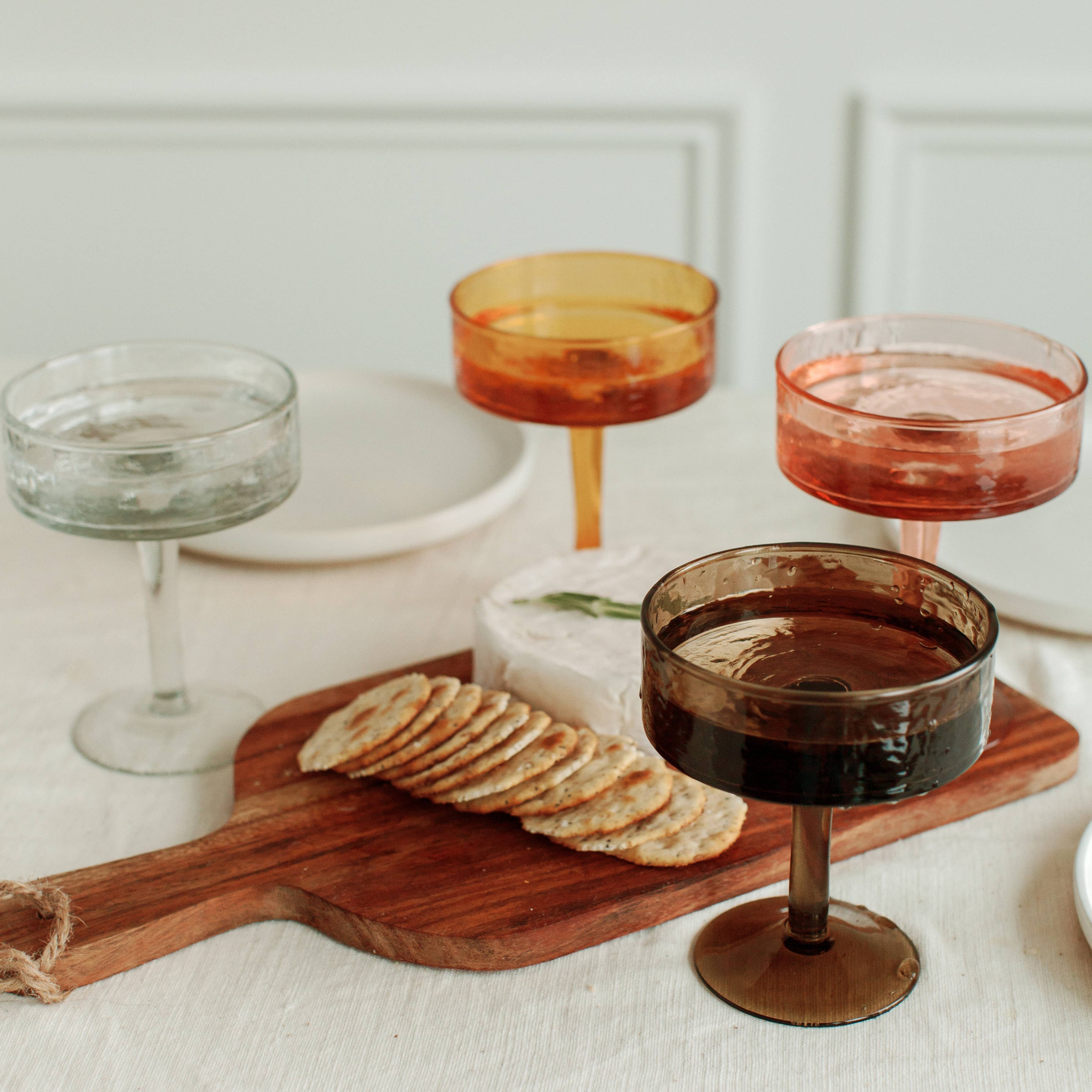 Cocktail glasses with different colored liquids on a wooden board with crackers.