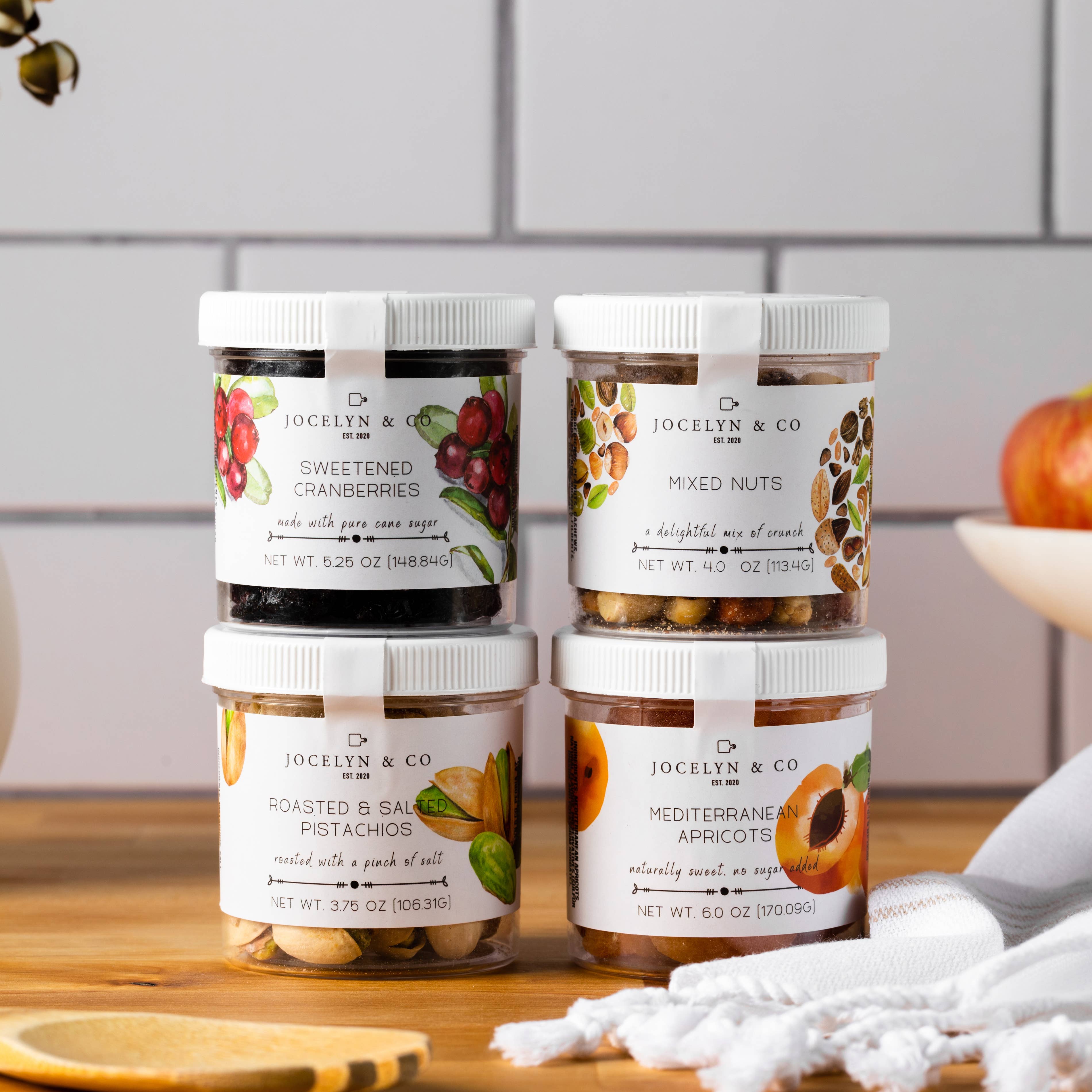Four jars of Jocelyn & Co. snacks on a wooden surface with a kitchen background.