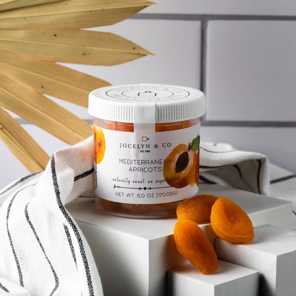 Jar of Jocelyn & Co. Mediterranean Apricots body scrub on a white surface with dried apricots and a towel.
