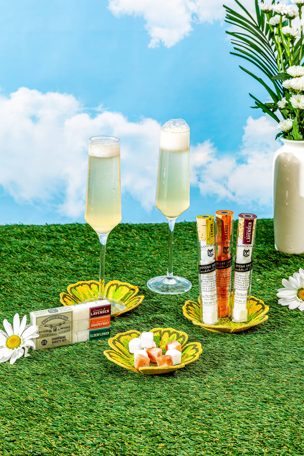 Two champagne flutes with a box of chocolates and a vase of flowers on grass with a blue sky.