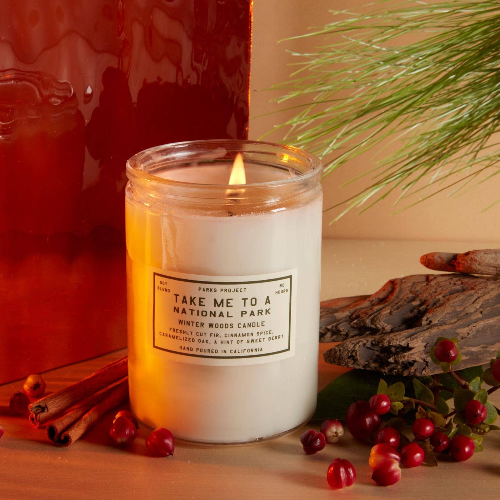 Lit candle in a glass jar with a label, surrounded by red berries and pine branches on a wooden surface.