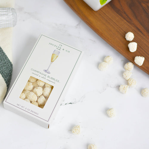 Box of champagne glass-shaped candies with a bottle of champagne and a plaid napkin on a white surface.