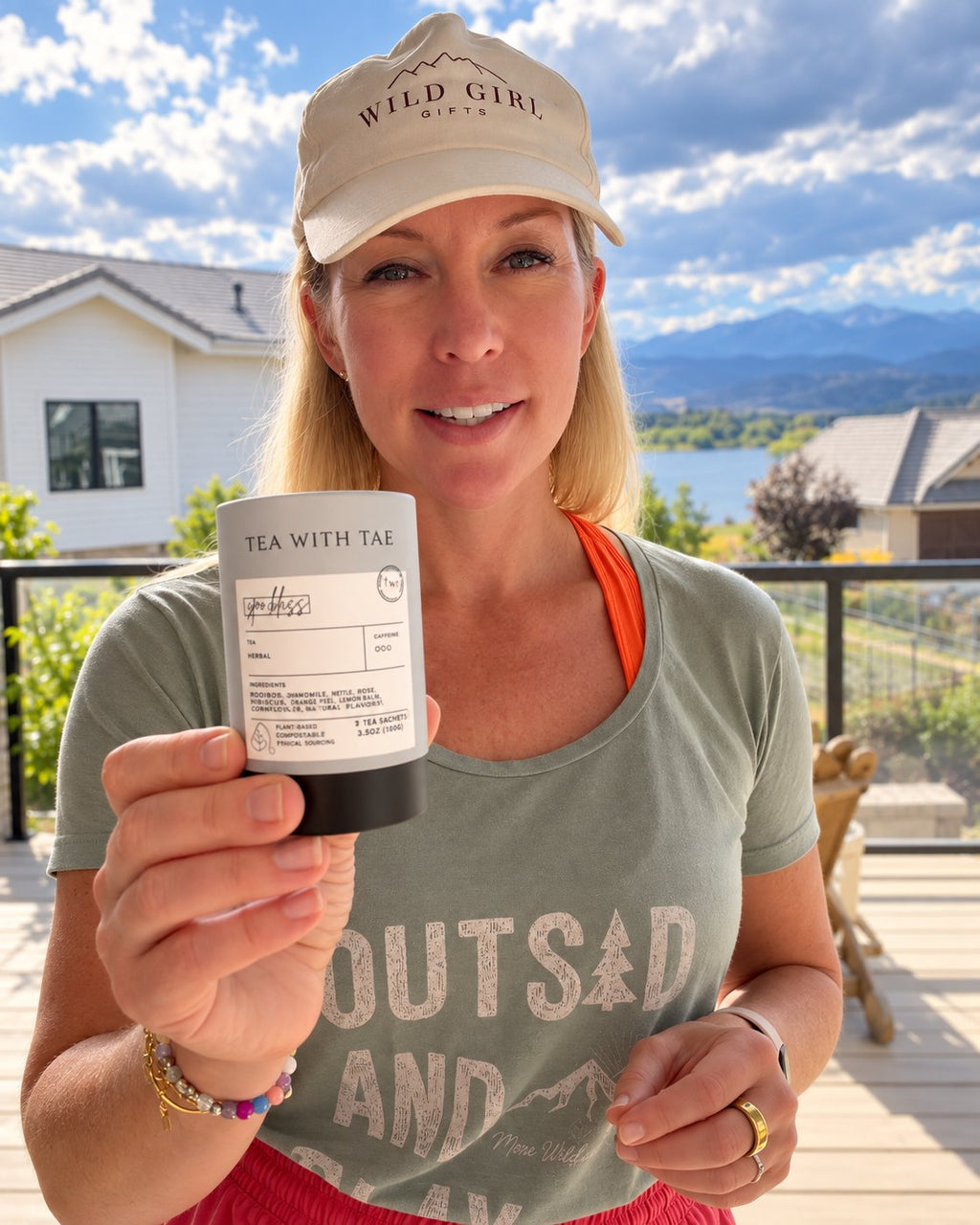 Kim in a Wild Girl Gifts cap holding a tin of Tea with Tae Spa Bliss herbal tea, with the Colorado foothills behind her.
