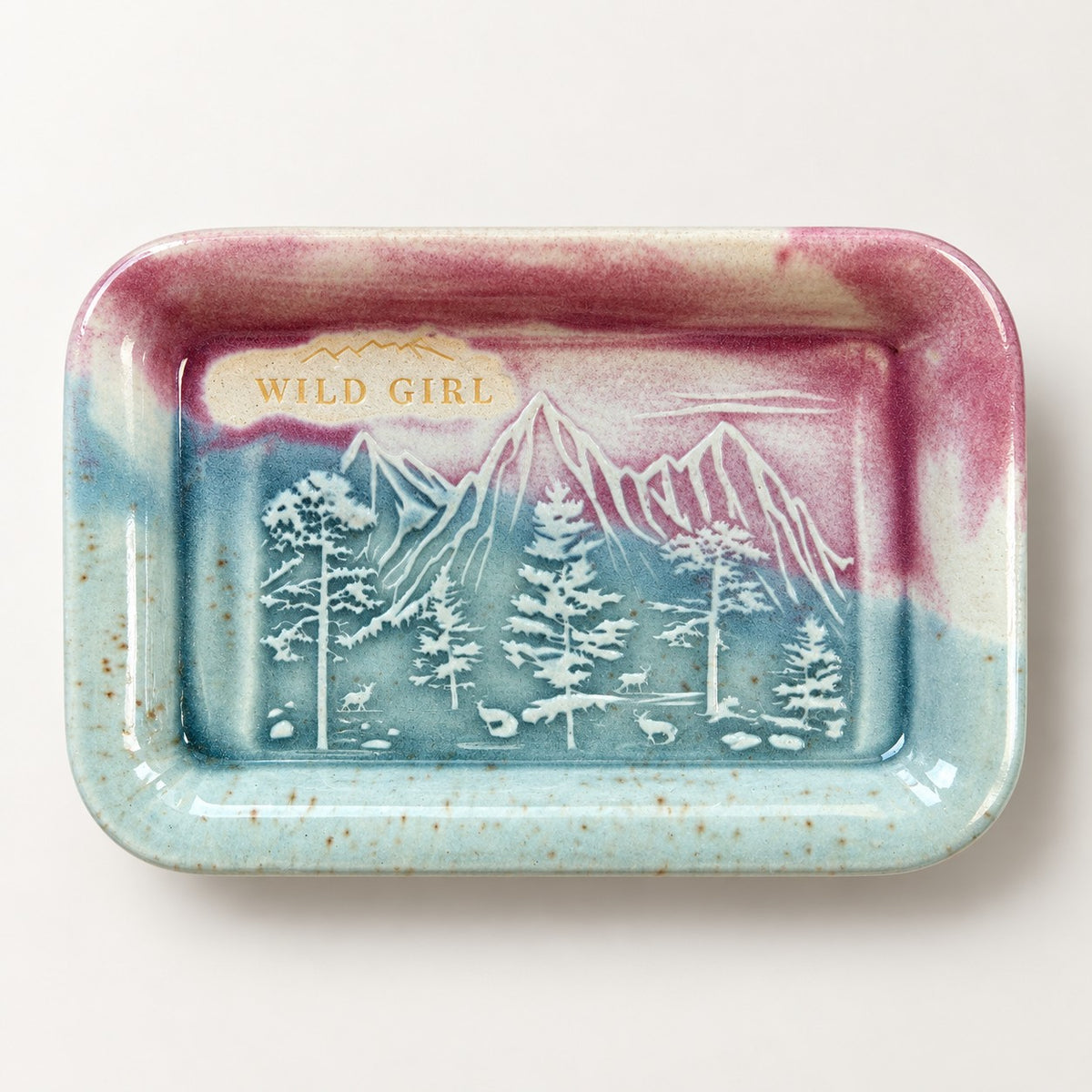 Wild Girl ceramic soap dish in the Sunrise Pink glaze, rose and alpine blue, with an embossed mountain range, pine trees and deer, and WILD GIRL stamped in gold.